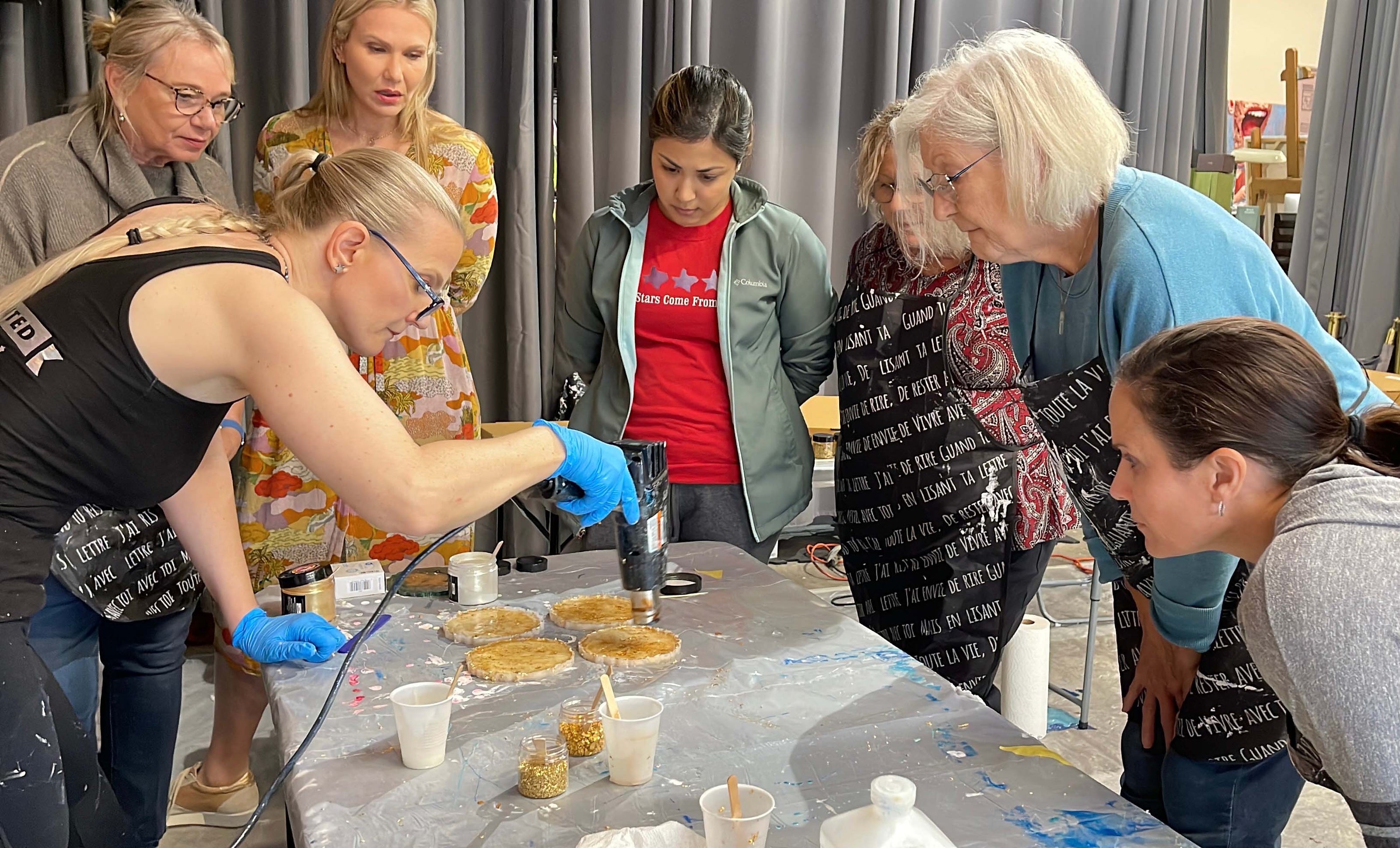 Anja Yamaji uses a heat gun to create gold resin coasters while workshop participants gather around to watch.