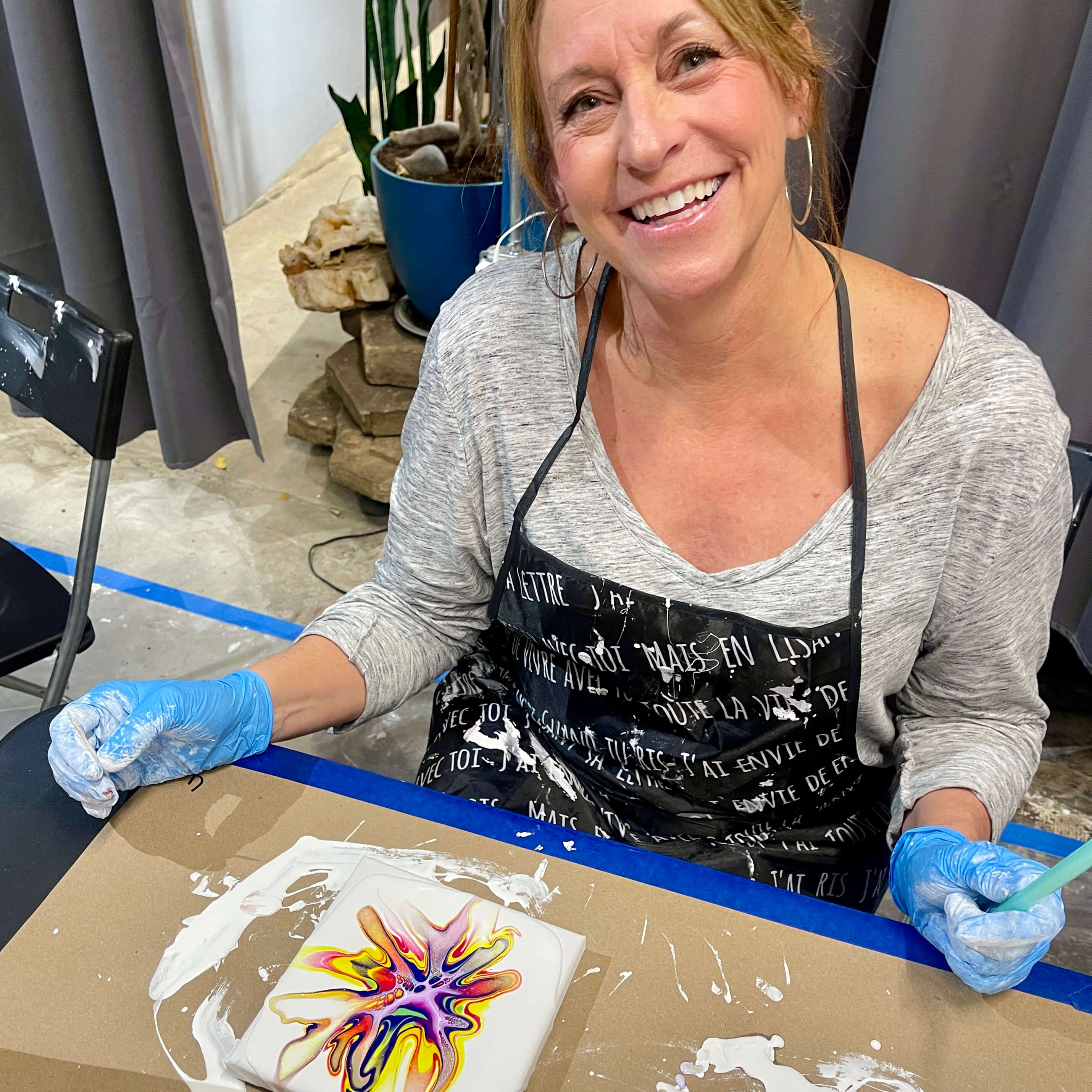 A student happily smiles at the camera with her acrylic flow painting displayed on the table in front of her.