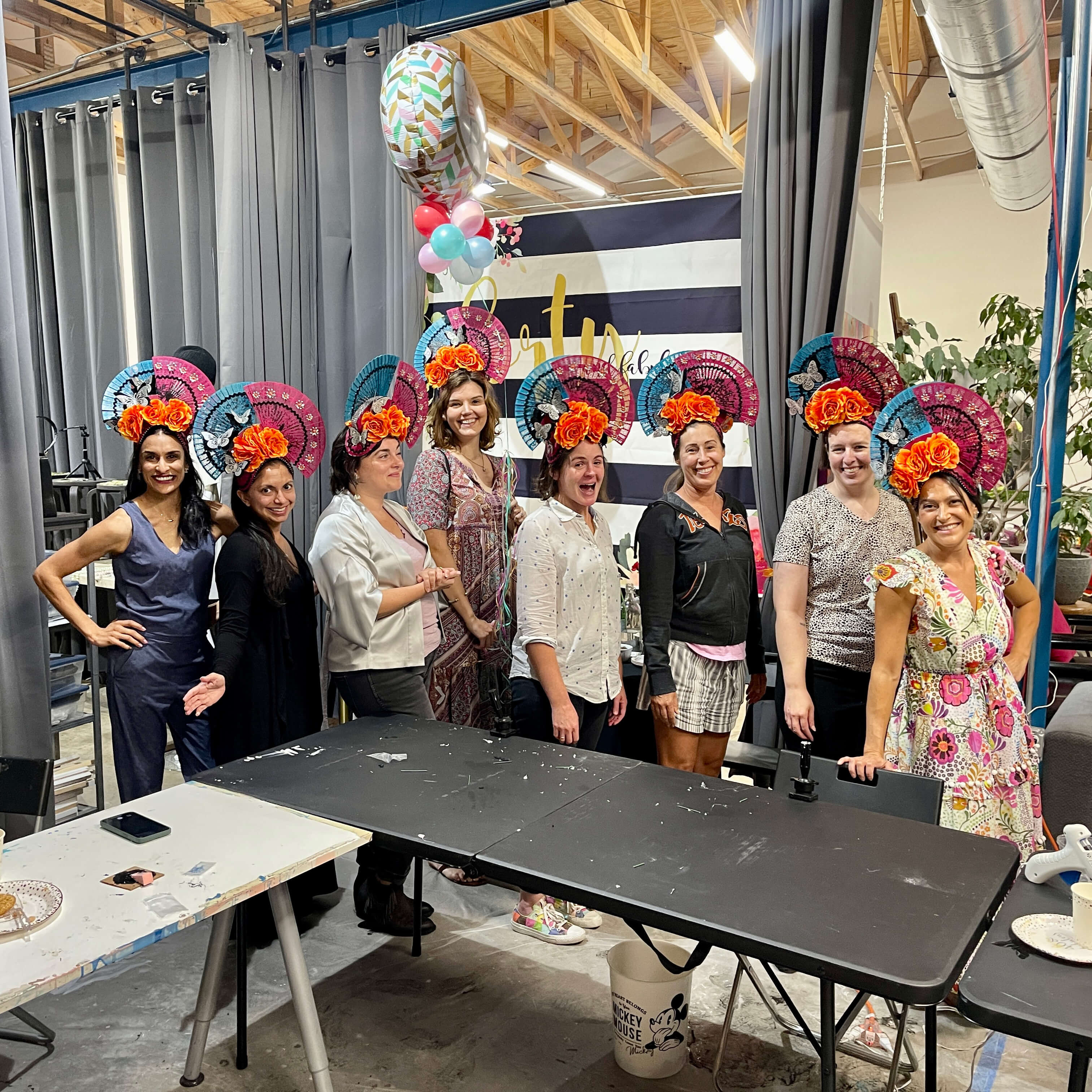 Group of eight ladies pose together, wearing the headdresses they created during their birthday celebration art experience.
