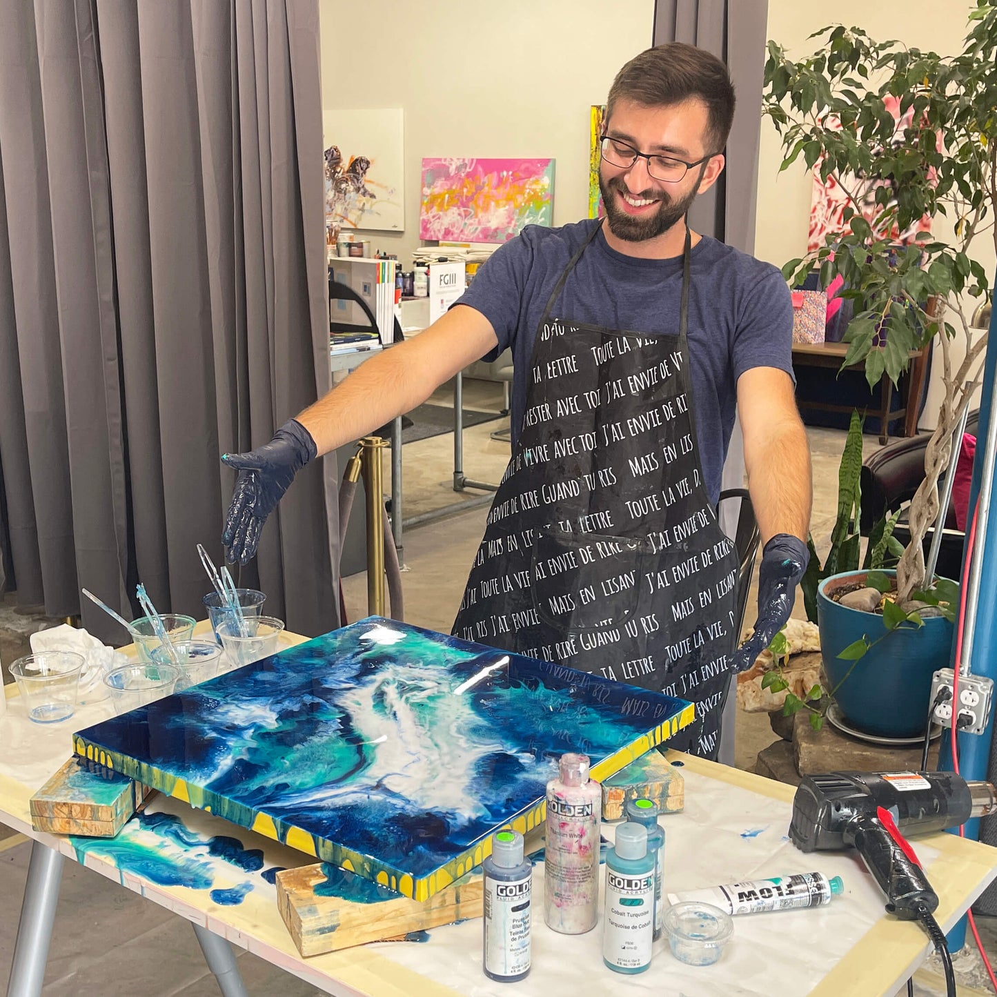A happy student stands behind his workstation, arms stretched out, proudly showing off his finished resin painting.