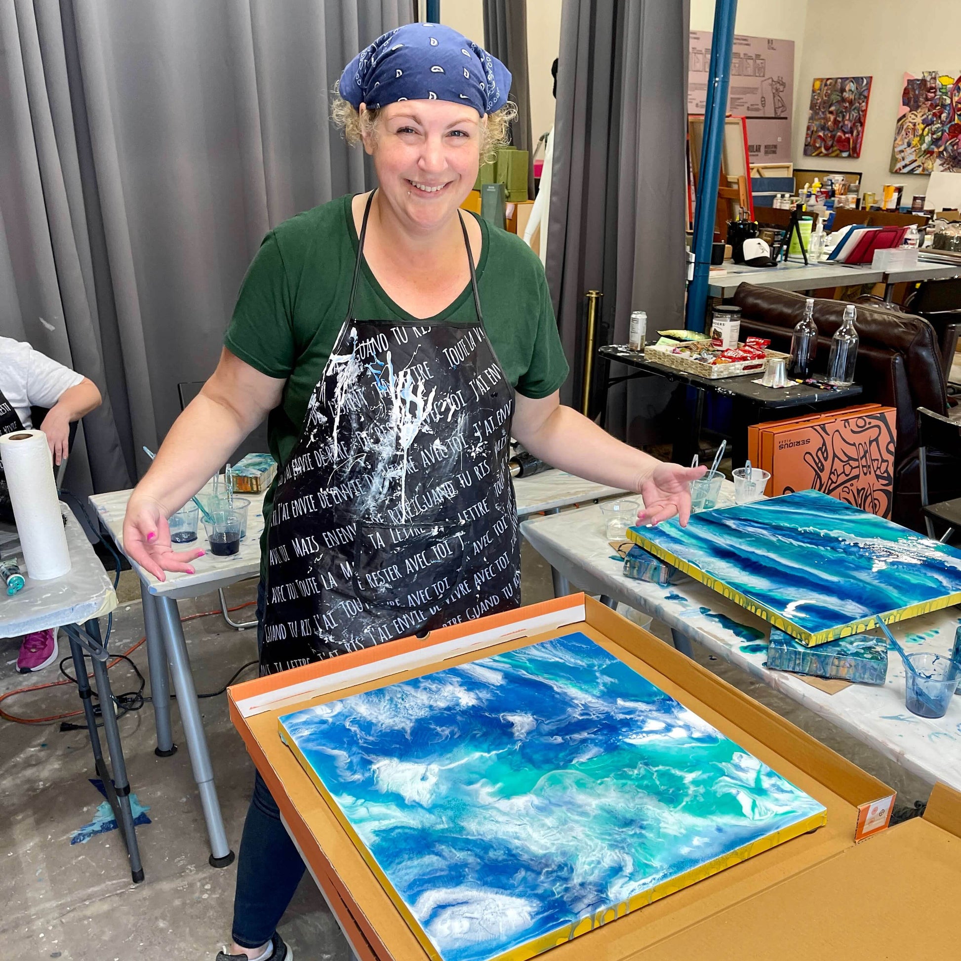 A smiling student poses for the camera with her finished ocean resin art painting laying in a transport box.