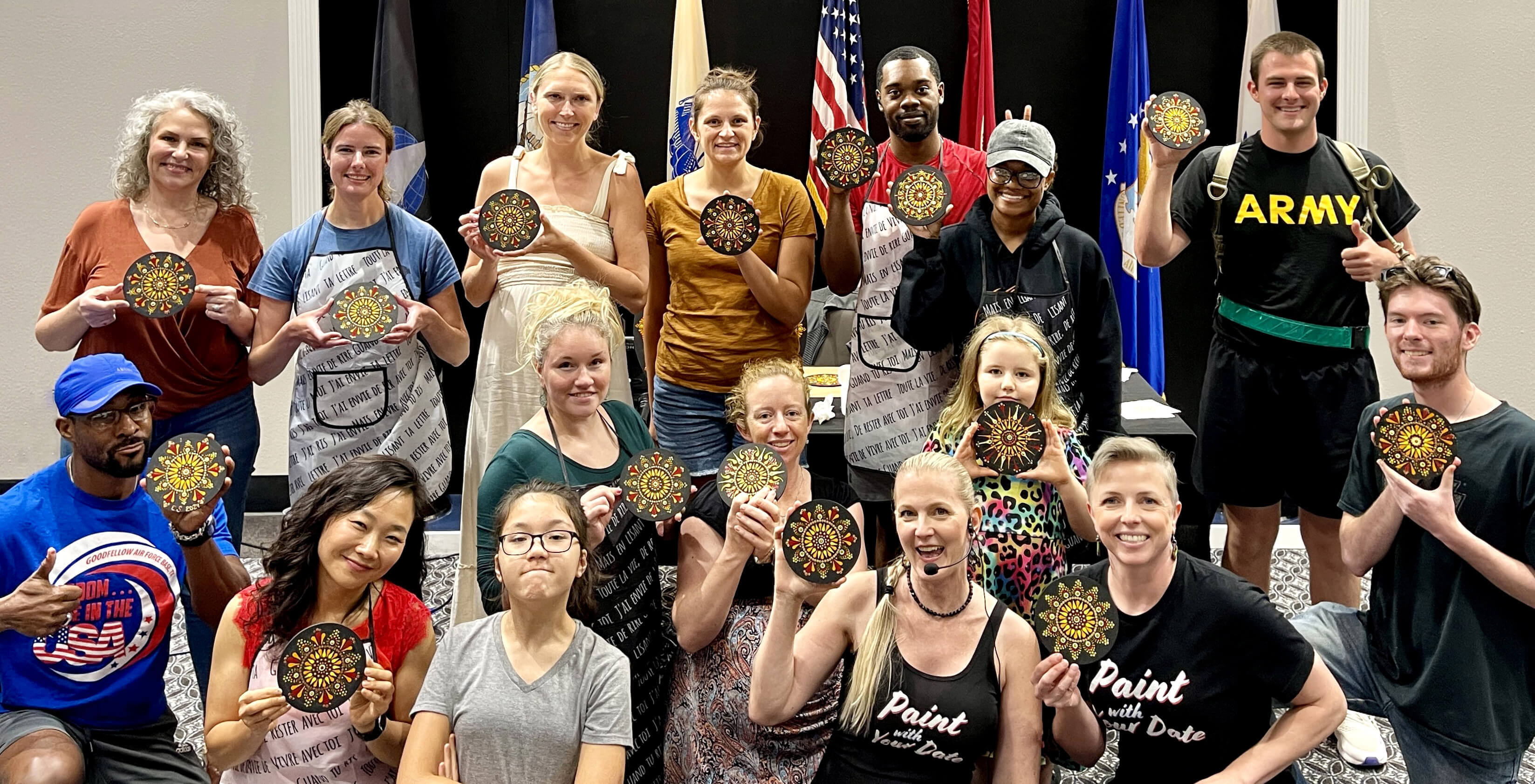 A large group of mandala art workshop participants proudly holding up their hand-painted mandalas after the experience.