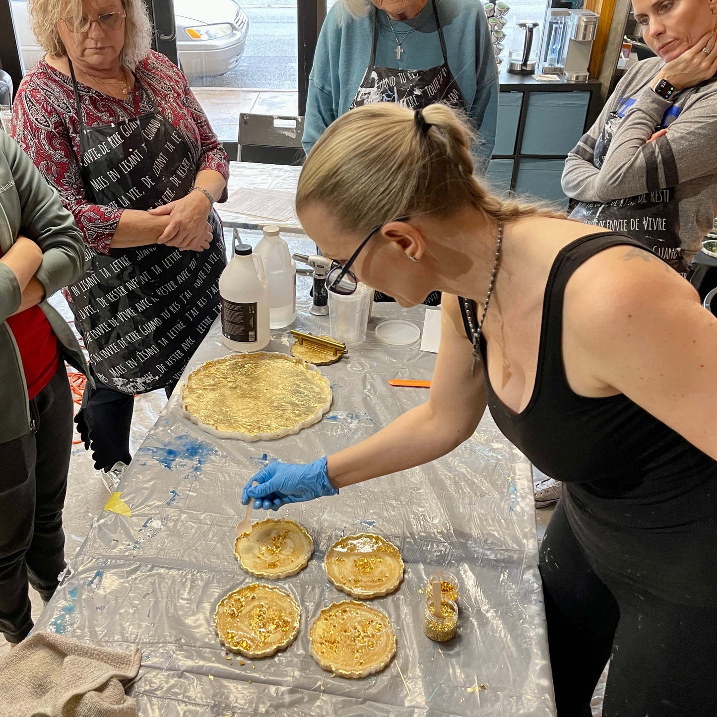 Anja Yamaji sprinkles crushed gold glass into her resin-filled silicone coaster molds as students watch attentively.