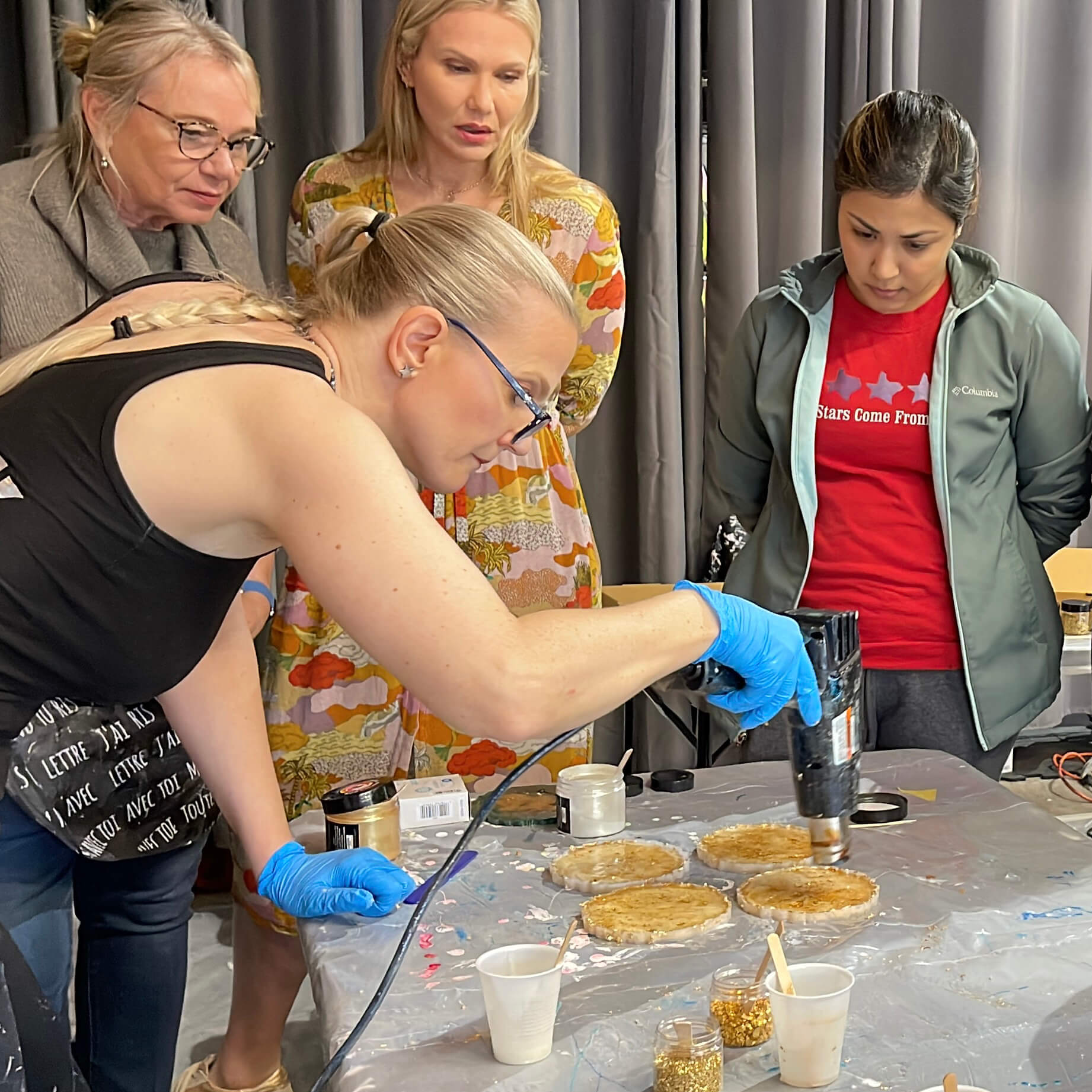 Anja Yamaji uses a heat gun to create gold resin coasters while workshop participants gather around to watch.