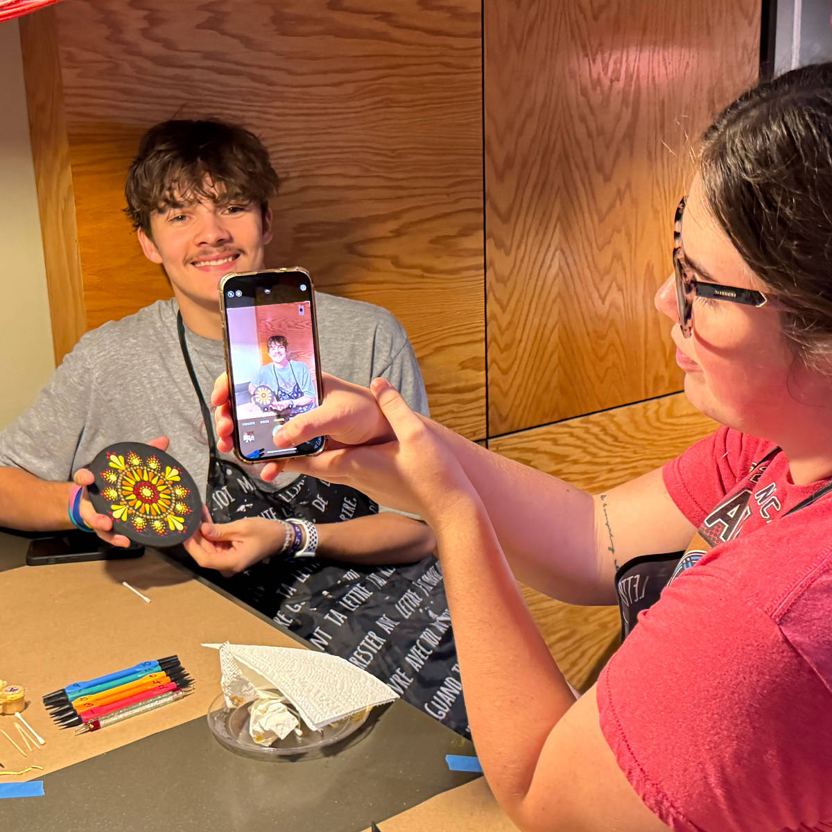 Two people in a casual setting, one holding a phone and showing his mandala painting to the other.