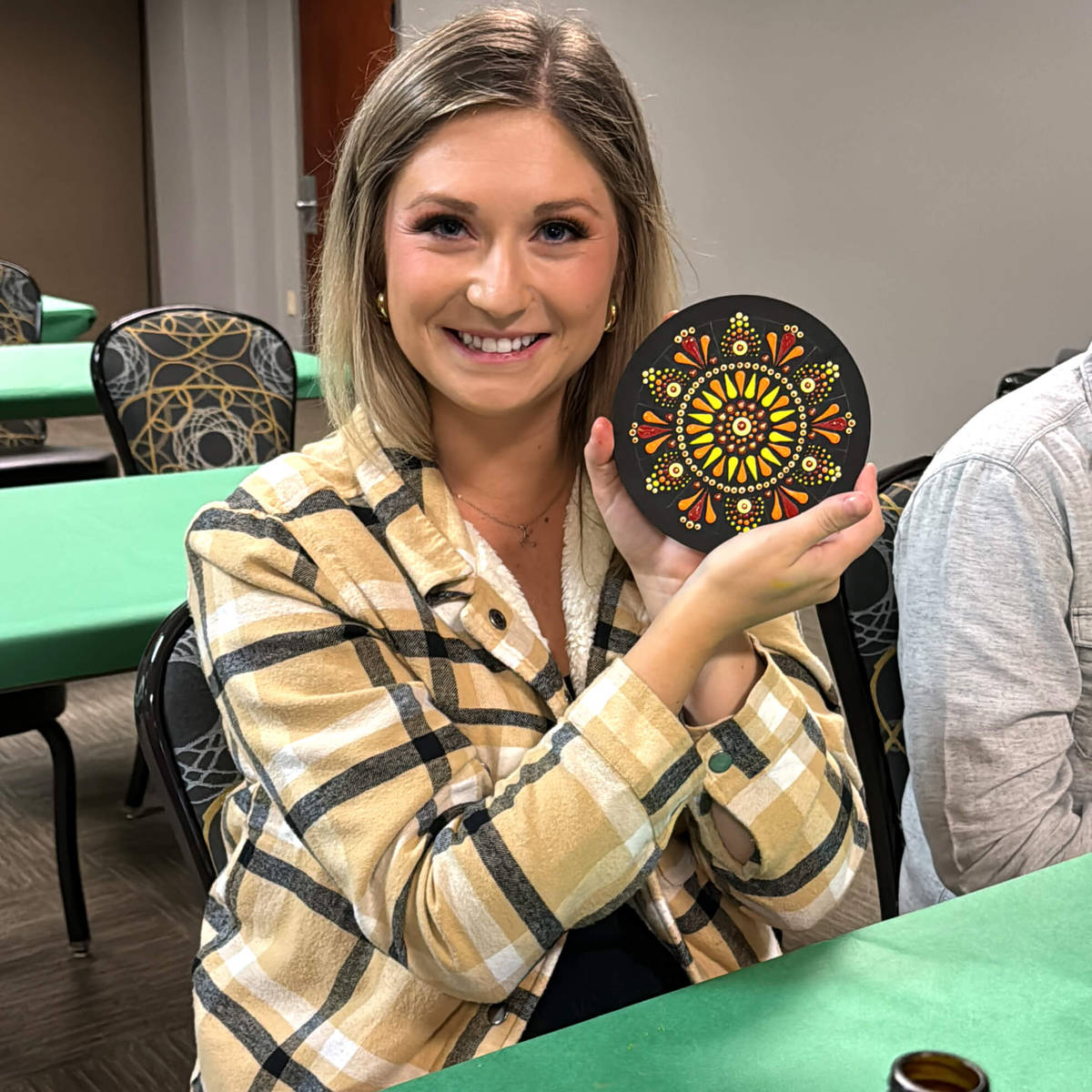 Woman holding a mandala painting in an indoor classroom setting.