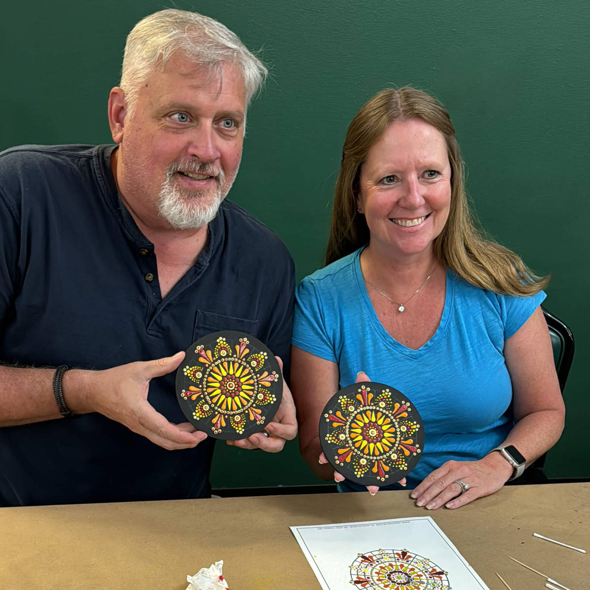 Man and woman holding mandala paintings at a table with a green background