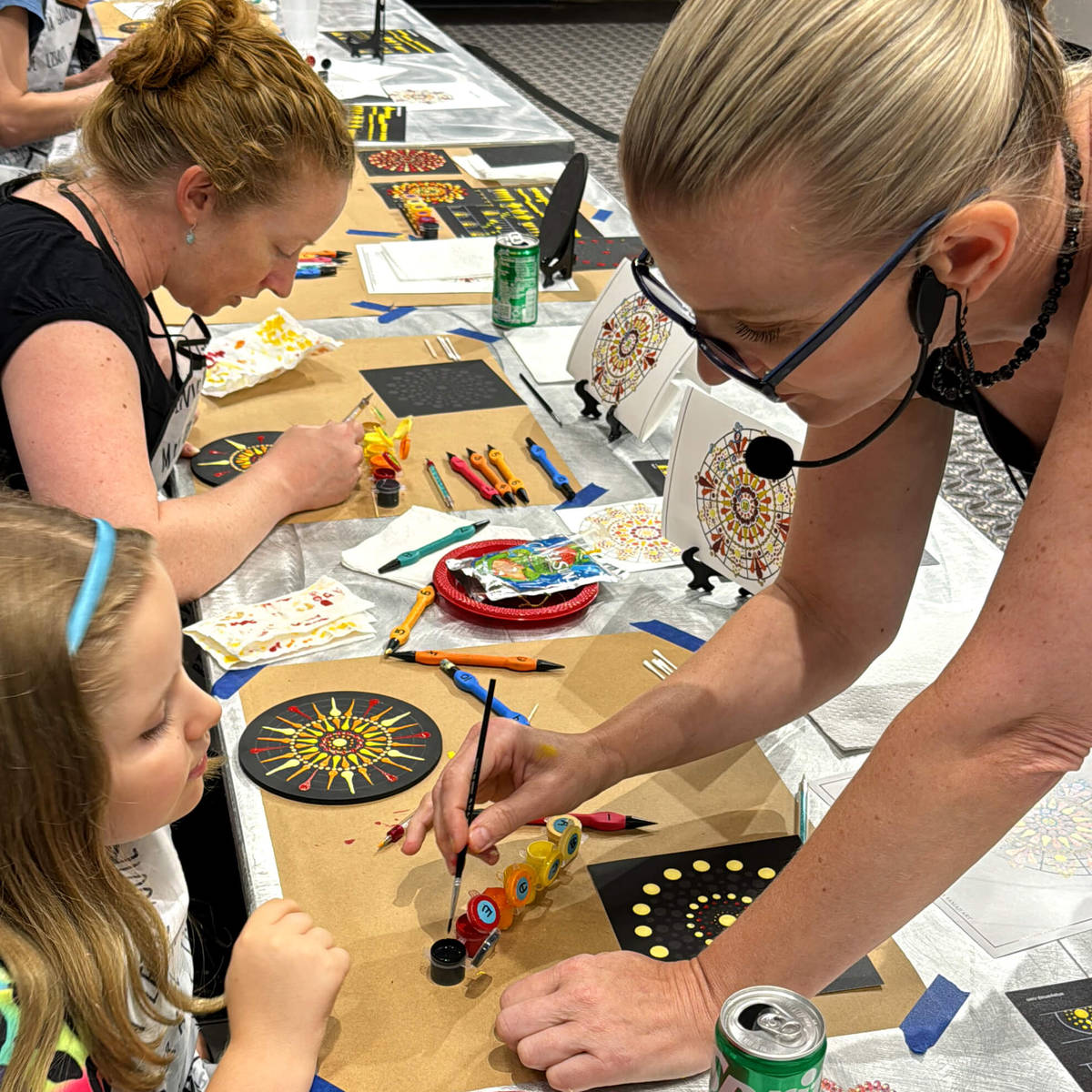 Two women and a child engaged in a mandala art workshop at a table with various materials.