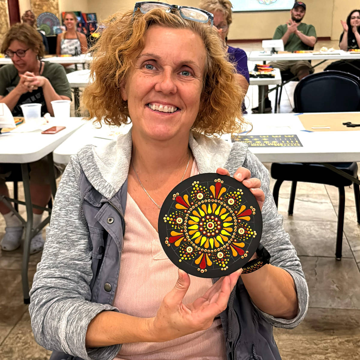 Woman holding a mandala painting in a casual indoor setting with people and tables in the background.
